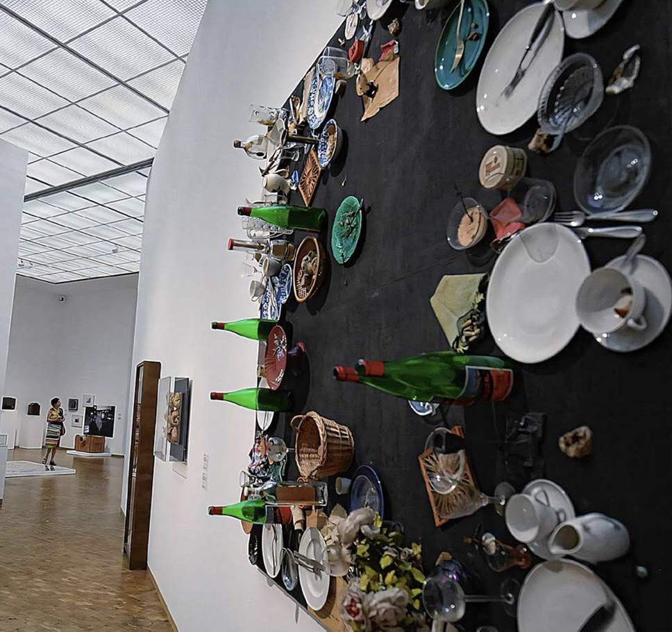 Museumsraum mit an der Wand befestigter Tischinstallation aus Tellern, Gläsern, Flaschen und Geschirr